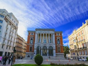 View of the Museo del Prado in Madrid, one of the most visited museums, with its grand neoclassical facade. The sky is vivid blue with wispy clouds. Surrounding the museum are historic buildings with detailed architecture. A group of people stand nearby on a sunny day, highlighting the lively urban scene.