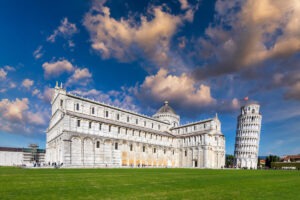 The image captures the Leaning Tower of Pisa and Pisa Cathedral during an Italian road trip under a dynamic sky with scattered clouds. The white marble buildings contrast beautifully with the vibrant green lawn, and warm sunlight highlights both structures amidst your Italy driving adventure.